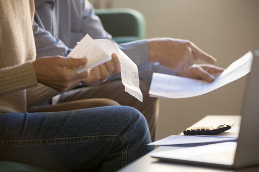 Elderly spouses sitting on couch at home planning budget check bills cheques, computer, documents and calculator on coffee table, close up cropped concept image, couple manage family expenses concept